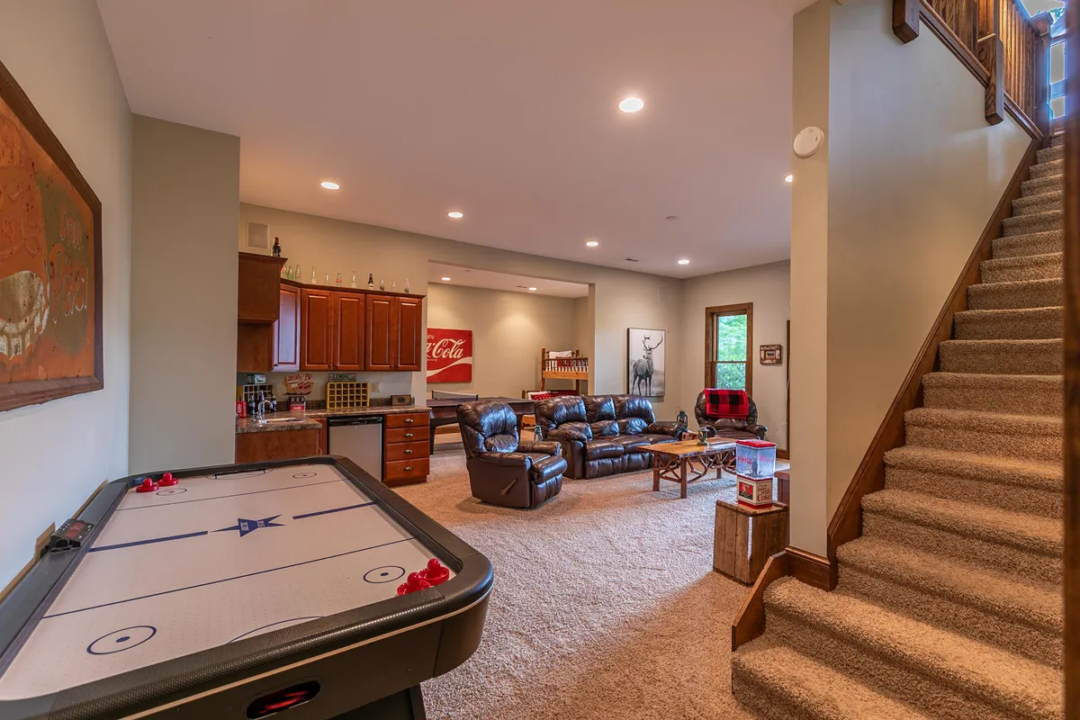 A relaxed basement game and media area with an air hockey table, plush leather couches, a kitchenette, and a staircase. The space is casual and perfect for both game nights and movie marathons, with ample room for group activities.