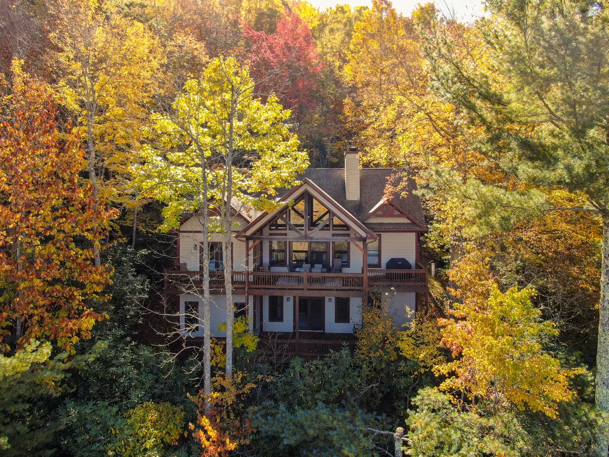 The house with a large upper balcony stretches out over a backdrop of vibrant autumn foliage, with wood and stone textures beautifully contrasting the surrounding natural scenery. The spacious balcony is clearly visible, ideal for gatherings and relaxation, while the landscape hints at serene outdoor living.