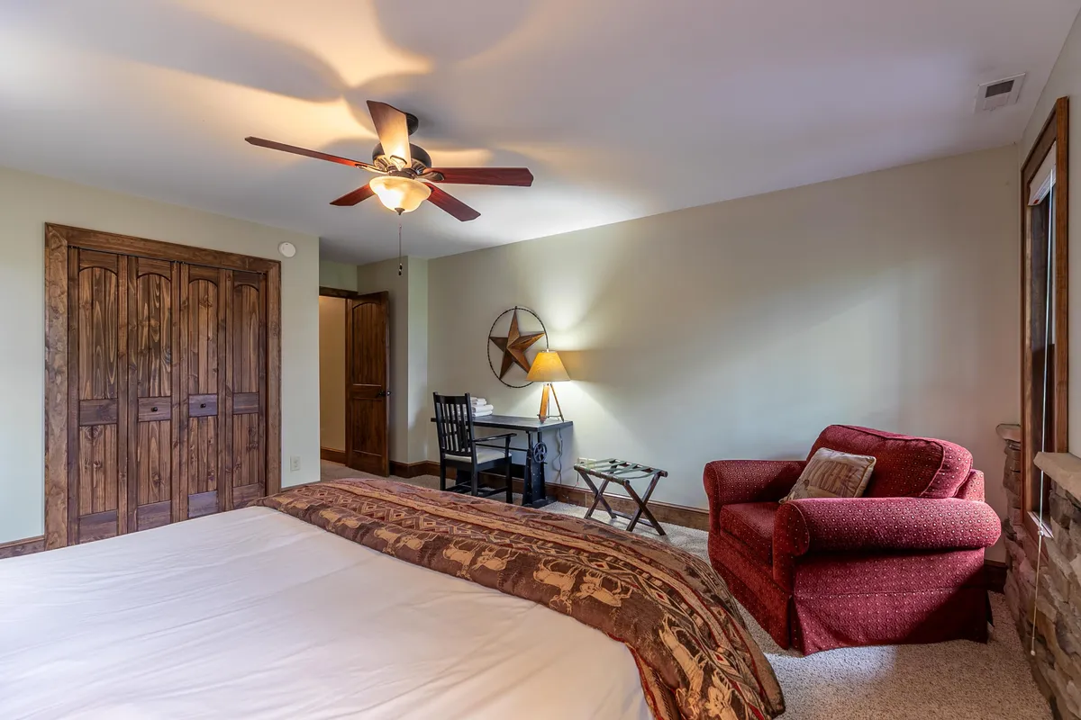 A spacious bedroom featuring a patterned bedspread, a red armchair, a wooden desk and chair, a ceiling fan, and a star decoration on the wall. The space is bathed in natural light, offering both relaxation and productivity.
