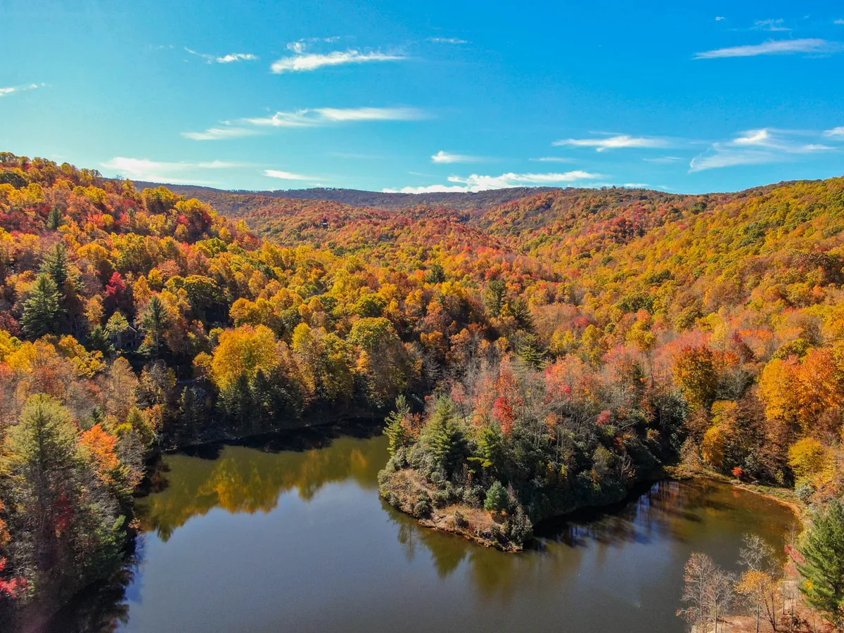 A breathtaking view of vibrant autumn foliage surrounding a serene, reflective body of water with rolling hills in the distance. The scene is alive with color, suggesting the beauty of hiking, fishing, or nature photography during the fall season.