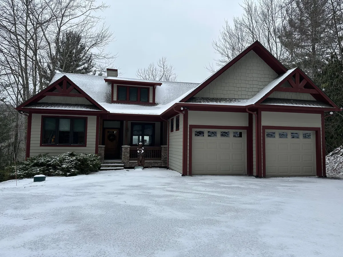The exterior of a charming mountain home dusted in snow, featuring a welcoming front porch, decorative gables, and garage doors. The scene depicts a cozy retreat perfect for winter activities and holiday gatherings, with snow blanketing the roof and surroundings.