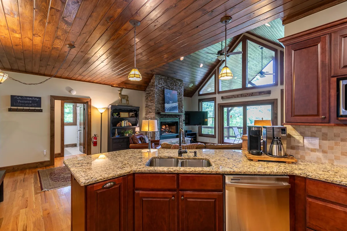 This image offers a view that blends the kitchen and living area, showcasing granite countertops, a five-burner gas stove, a kitchen island, the stone fireplace, and large windows. The hardwood floors and wooden ceiling enhance the cohesive, upscale rustic feel.