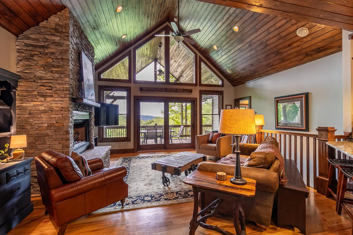This image presents a spacious living room with a prominent stone fireplace, wooden vaulted ceilings, large windows letting in abundant natural light, comfortable seating, and a central coffee table. The atmosphere is both warm and inviting, with ample space for gathering and relaxing.