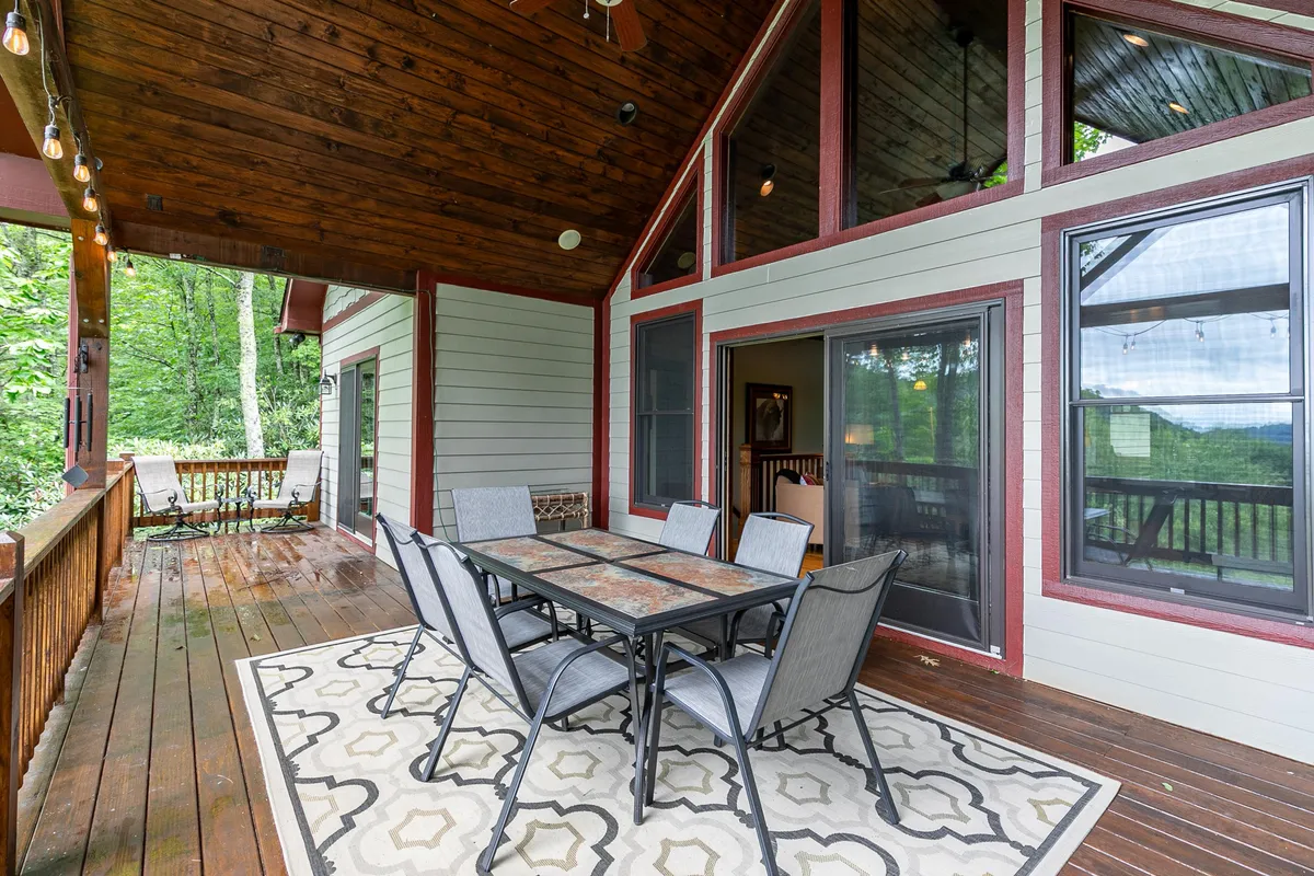 A welcoming outdoor dining area sits on a covered deck with a sturdy wooden ceiling and large windows reflecting the greenery outside. A table set for a meal is surrounded by comfortable chairs, creating a perfect space for sunset dinners or morning coffee amidst nature.