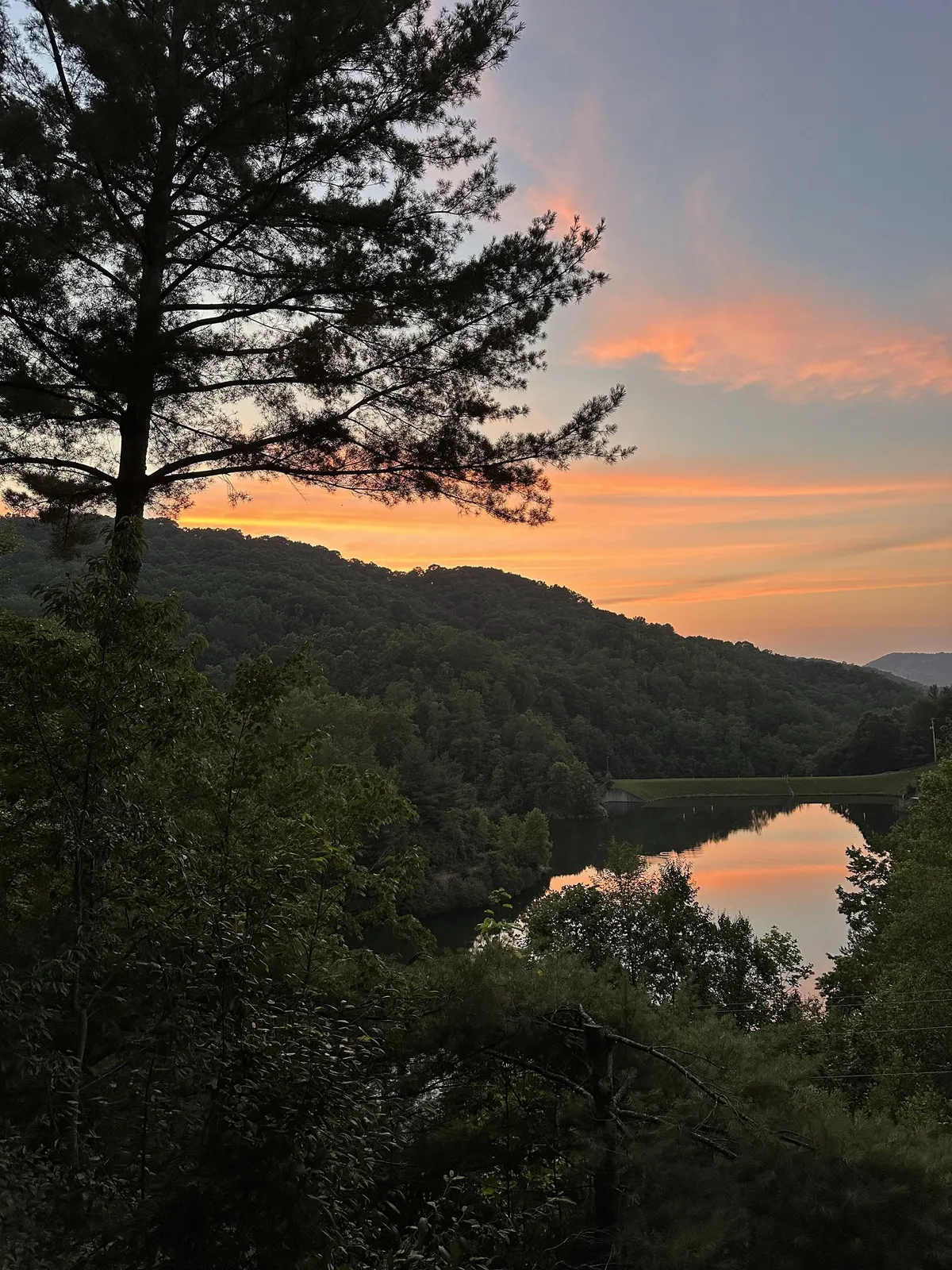 This image captures a stunning sunset over the mountains, with the water reflecting the vibrant sky and the silhouettes of trees framing the scene. The tranquil atmosphere evokes the serenity of summer evenings spent outdoors.