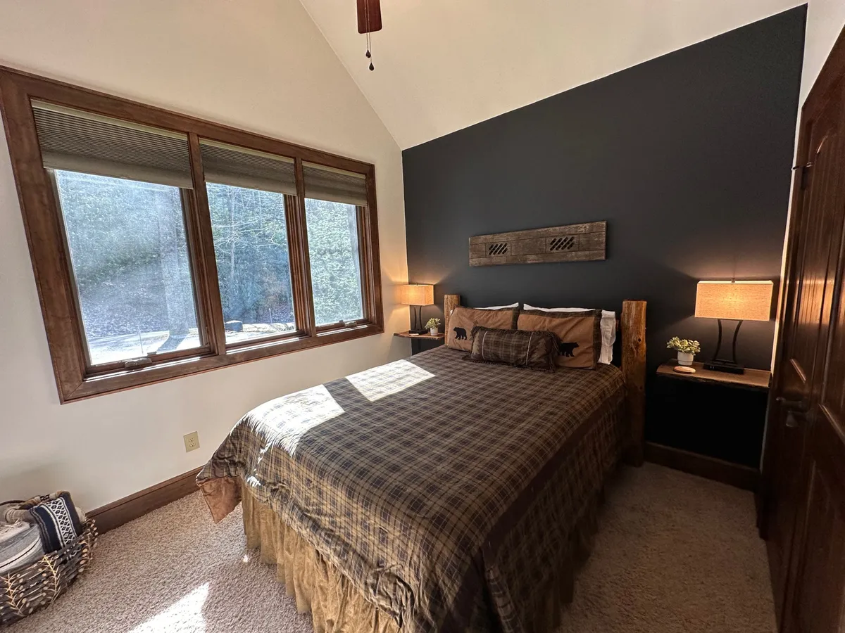 A serene bedroom with plaid bedding, large windows offering abundant natural light, and a nightstand with a lamp, creating a comfortable and peaceful ambiance ideal for relaxation or reading.