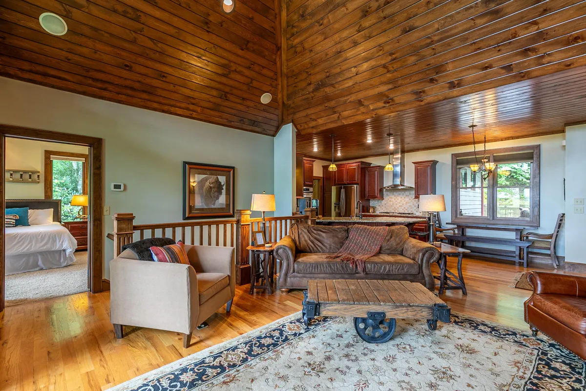 The image highlights a large leather sofa positioned under wooden ceilings, with a view of the adjacent dining area and a decorative rug anchoring the space. The overall mood is cozy and conducive to social gatherings.